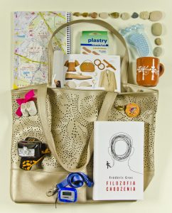 Zdjęcie torby z akcesoriami: książką Filozofia chodzenia oraz olanem miasta, kubkiem, kamykami, plastrami, latarką czołówką, odznakami.