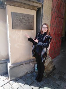 uśmiechnięta kobieta w okularach pokazuje kamienną tablicę na fasadzie budynku; na tablicy napis "Narodni knihovna Ceske Republiky"