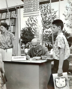 Stare zdjęcie. Mężczyzna podpisuje książkę, obok stoi chłopiec z torbą w ręku.
