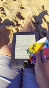 Zdjęcie trzymanego w ręcze czytnika ebooków oraz maseczki, w tle wyciągnięte na piasku nogi.