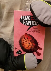 Zdjęcie okładki książki trzymanej przez człowieka przebranego za maskotkę. Na różowej okładce ryba rozdymka w kolorze pomarańczowo-czerwonym.