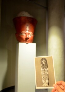 Zdjęcie, na pierwszym planie pocztówka z drewnianym, egipskim sarkofagiem, w tle rzeźba królowej Hatszepsut w wolnym dostępie BUW.