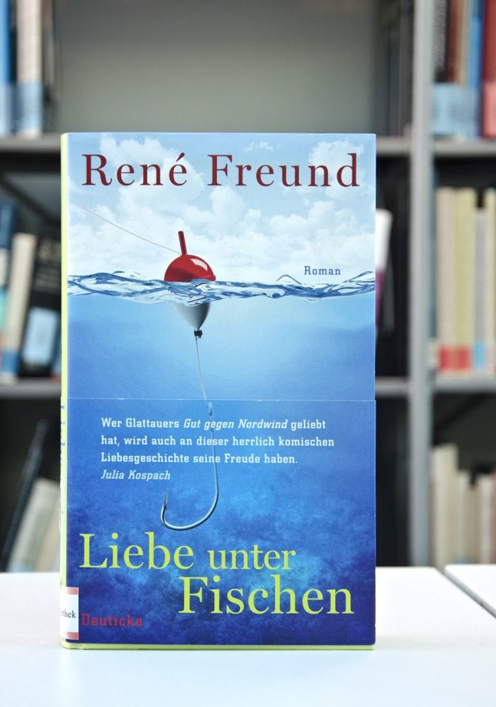 Książka pt. Liebe unter Fischen na tle regału z książkami. Okładka przedstawia spławik widziany z poziomu wody.