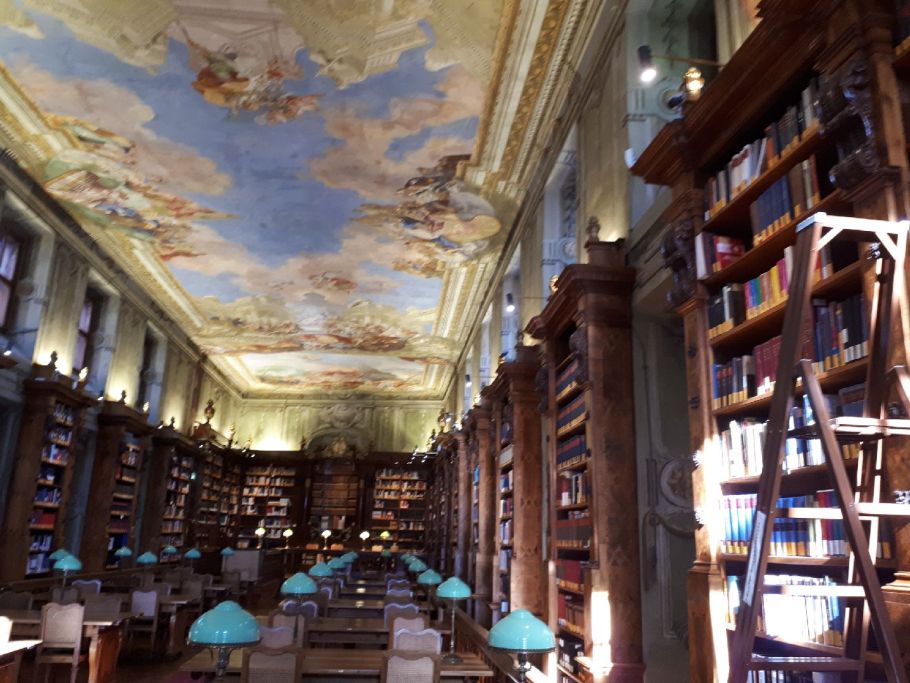 Zdjęcie zabytkowej czytelni bibliotecznej z drewnianymi regałami, stołami doświetlanymi lampami z zielonymi kloszami. Na suficie widać fresk.