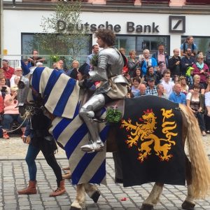 Zdjęcie ubranego w zbroję rycerza siedzącego na koniu, którego prowadzi giermek. W tle widać publiczność oglądającą paradę.