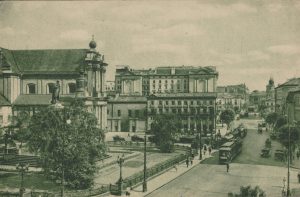 Zdjęcie międzywojennego wyglądu Krakowskiego Przedmieścia, Na pierwszym planie pomnik Mickiewicza, za nim Kościół oraz dzisiejszy Pałac Prezydencki. Na ulicy widać samochody oraz tramwaje.