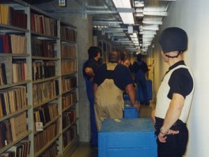 Zdjęcie przeprowadzki zbiorów bibliotecznych. Widoczni mężczyźni przewożący kontenery transportowe oraz konwojent w hełmie i w kamizelce kuloodpornej.