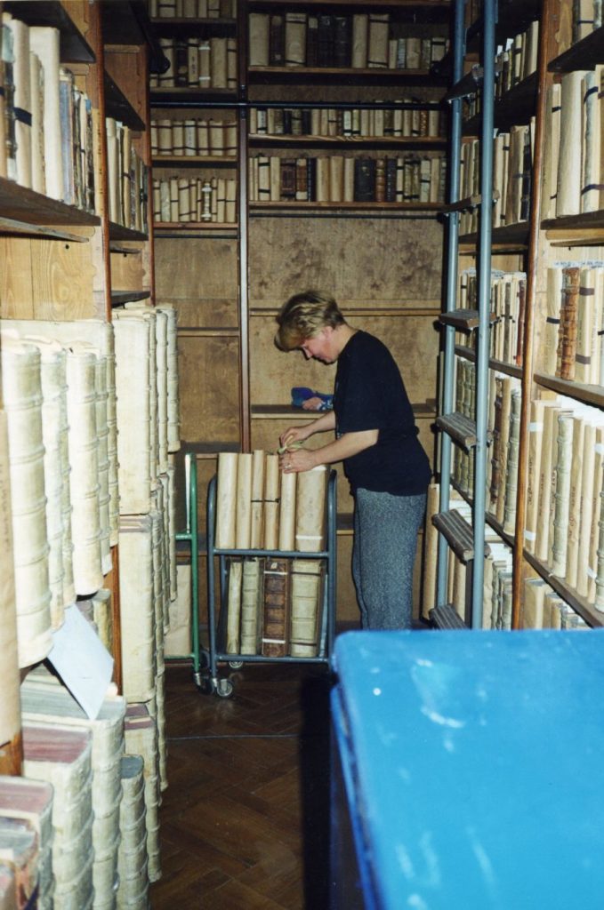 Zdjęcie kobiety przenoszącej duże księgo na wózek biblioteczny. Wokół zastawione księgozbiorem drewniane regały biblioteczne.