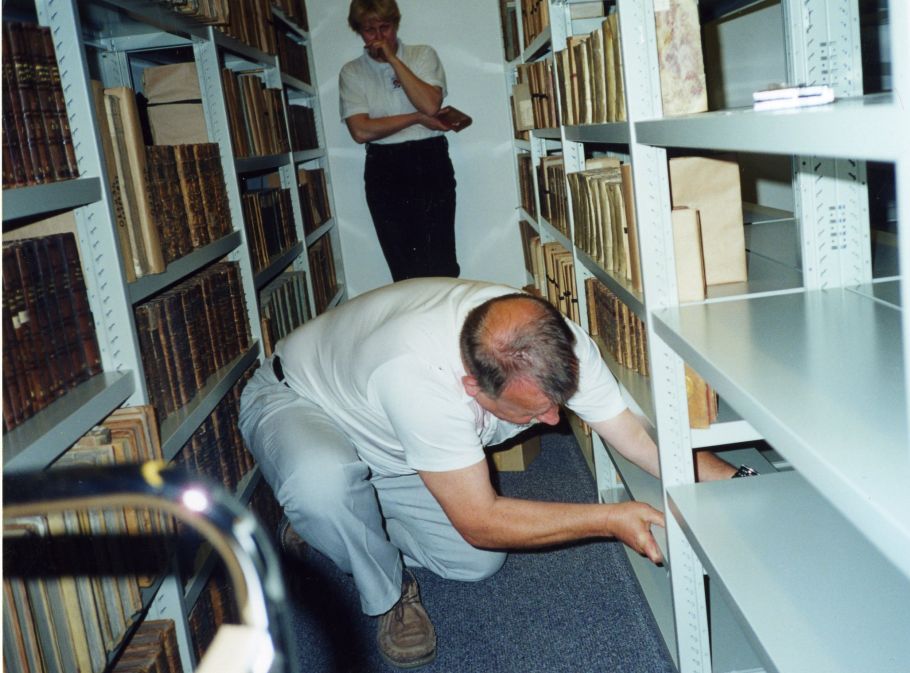Na zdjęciu widoczny mężczyzna regulujący wysokość półki w regale w magazynie bibliotecznym. W tle zapełnione woluminami regały.
