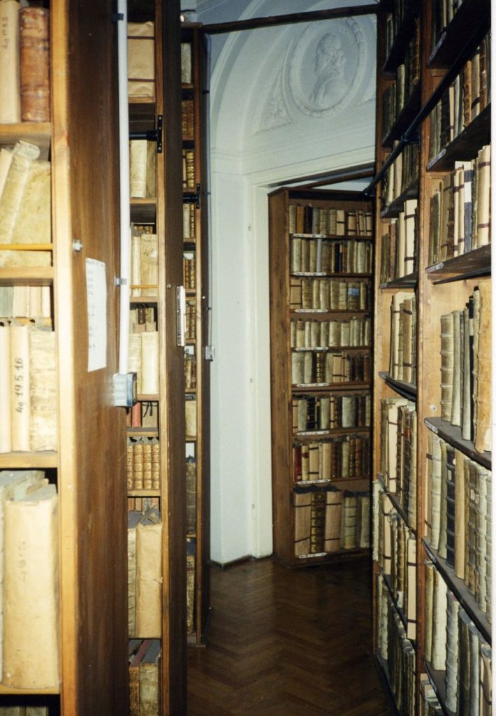 Zdjęcie magazynu bibliotecznego z drewnianymi regałami pełnymi książek. W dalszym planie widoczna płaskorzeźba nad otworem drzwiowym.
