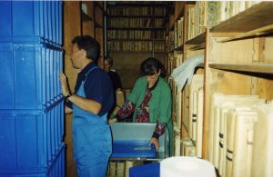 Zdjęcie Trzech osób pakujących książki w folię bąbelkową a następnie do kontenerów transportowych w magazynie bibliotecznym zastawionym już częściowo pustymi drewnianymi regałami.
