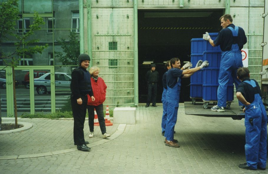 Zdjęcie wyładunku z ciężarówki kontenerów transportowych przed bramą wjazdową do BUWu na Powiślu. Widoczni pracownicy przewożący kontenery oraz konwojenci i pracownica biblioteki.