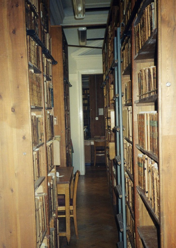 Zdjęcie magazynu bibliotecznego. Widać przejście między drewnianymi regałami zastawionymi książkami.