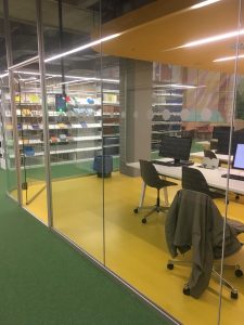 Zdjęcie przeszklonej czytelni bibliotecznej. W środku stoły, a na nich klawiatury i monitory komputerowe.