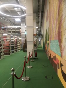 Zdjęcie biblioteki. Po lewej widać regały zastawione oprawionymi woluminami. U góry okrągły nowoczesny żyrandol. Po prawej ściana, na której widać kolorowe malowidło.