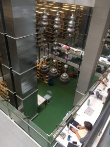 Zdjęcie nowoczesnej przestrzeni bibliotecznej. Widać regały z książkami, filary obudowane srebrnym metalem, szklane barierki oraz czytelników pracujących przy stołach. Z sufitu zwieszają się długie ozdobne metalowe żyrandole.