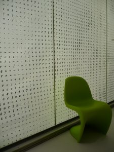 Zdjęcie zielonego fotela na tle mlecznobiałej szyby z literami, cyframi oraz innymi symbolami.