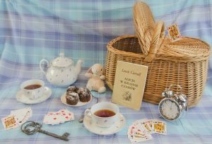 Na zdjęciu koszyk piknikowy, karta Dama Kier, budzik, rozłożona talia kart, dwie filiżanki herbaty, klucz, talerzyk z trzema muffinkami, imbryk, maskotka królik i książka "Alicja w krainie czarów".