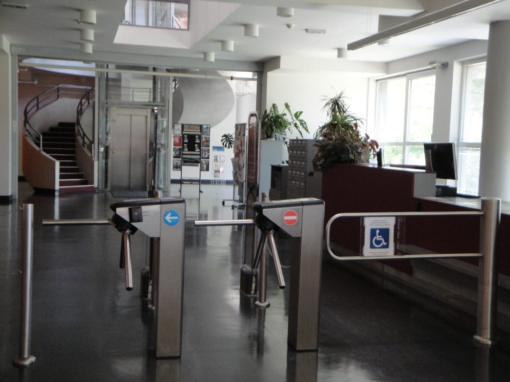Zdjęcie wejście do biblioteki. Widać kołowrotkowe bramki, oddzielne dla wchodzących i wychodzących oraz przejście dla osób niepełnosprawnych. W tle stanowisko biblioteczne, winda i okalające ją schody prowadzące w górę.