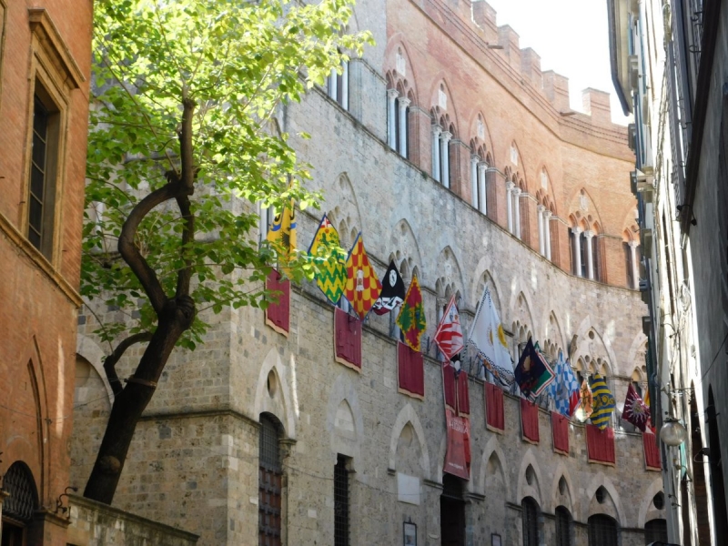 Ulica w Sienie. Średniowieczna kamienica z wywieszonymi flagami. Obok kamienicy dzrzewo.