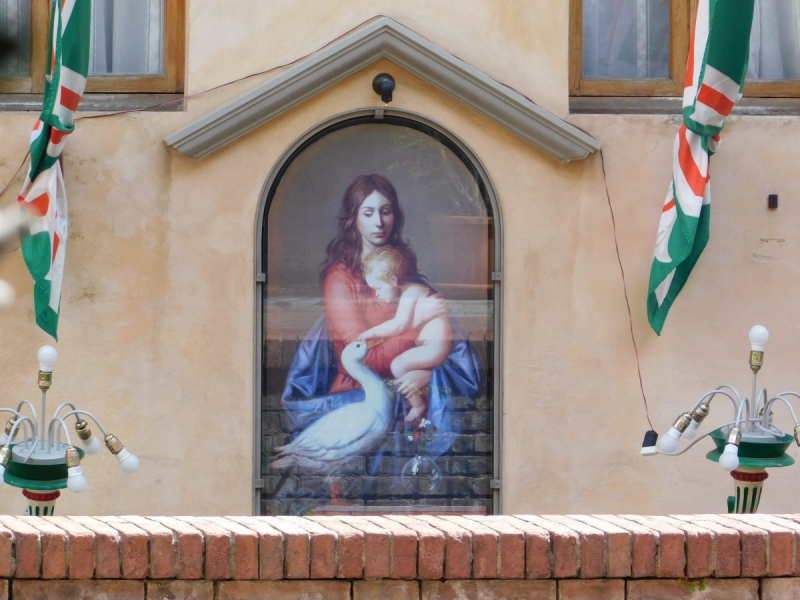 Kapliczka na murze domu. W kapliczce obraz kobiety dzieckiem głaszczącym kaczkę.