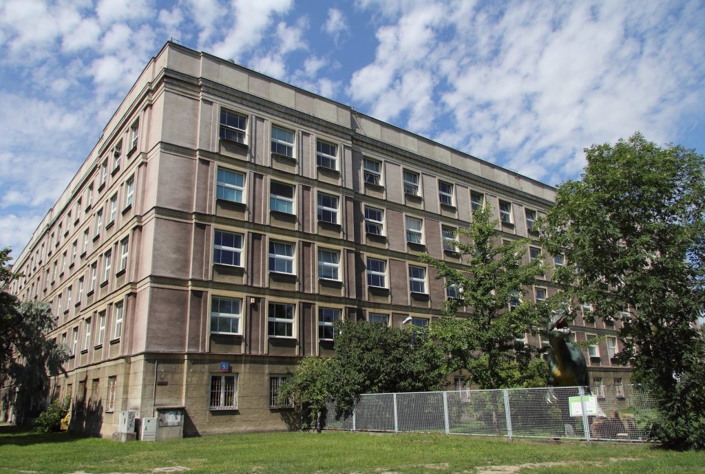 Zdjęcie budynku Wydziału Geologii UW. Na zdjęciu róg czteropiętrowego budynku i metalowe ogrodzenie (siatka) za ogrodzeniem model Tyranozaura.