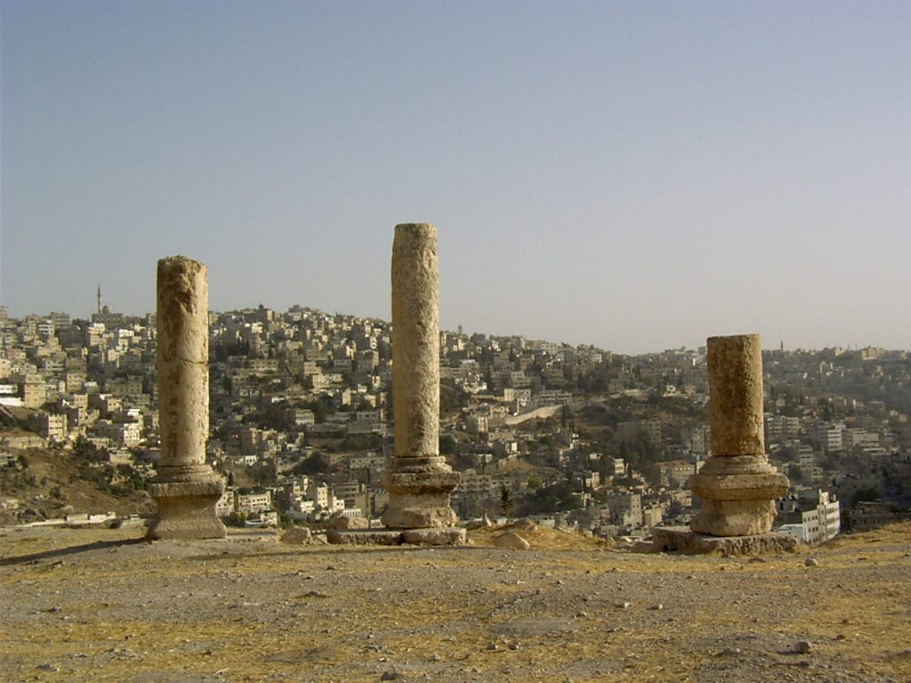 Na pierwszym planie trzy fragmenty kolumn, na dalszym planie miasto na wzgórzach