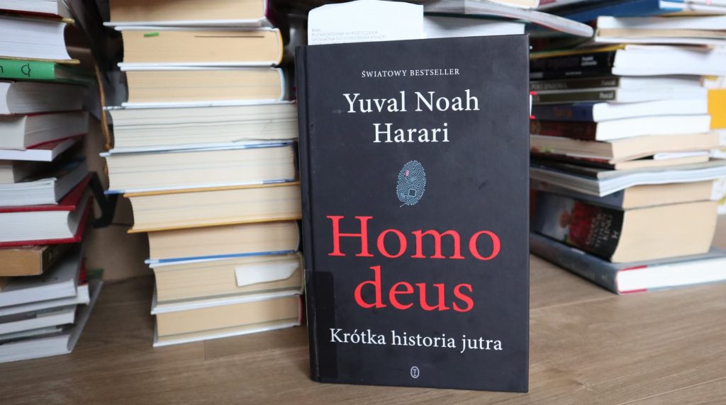 Czarna okładka publikacja Yuval Noah Harari Homo deus. W tle sterty książek czekających na opracowanie.