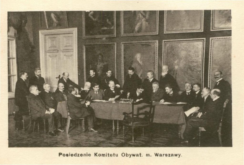 Stare czarno-białe zdjęcie przedstawiające mężczyzn siedzących przy stole. Na ścianach portrety wiszące w dwóch rzędach.