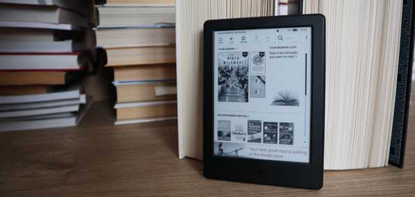 czytnik Kindla z wyświetlaczem startowym na tle stosów książek papierowych ułożonych na podłodze drewnianej