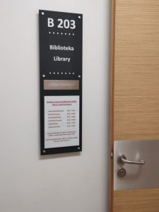 Zdjęcie czarnej tabliczki informacyjnej w ułożeniu pionowym. Na niech napis B 203, Biblioteka, poniżej zapis a w alfabecie Braille'a oraz godziny pracy.