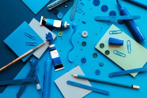 Na niebieskim tle rozrzucone niebieskie ołówki ,flamastry, tubki z farbą, pędzle, guziki, karteczki i spinacze.