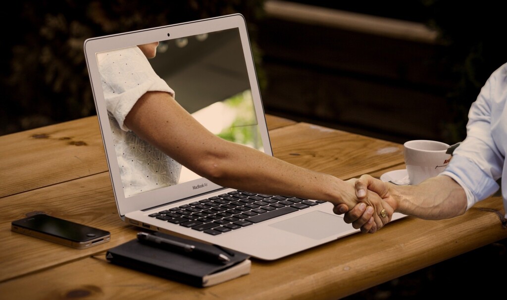 Ręka wychodząca z ekranu laptopa ściskająca rękę użytkownika laptopa