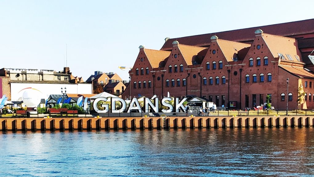 Zdjęcie napisu Gdańsk, na pierwszym planie woda, za napisem zabytkowe budynki