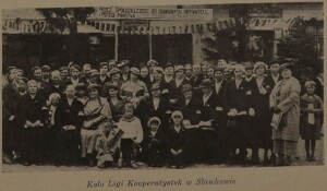 Zdjęcie grupy kobiet i dziewczynek poozujących na tle budynku ozdobionego hasłem: przez spółdzielczość do dobrobytu obywateli i potęgi państwa [niewyraźny wyraz] wszyscy do spółdzielni! Zdjęcie podpisane: Koło Ligi Kooperatystek w Sławkowie.