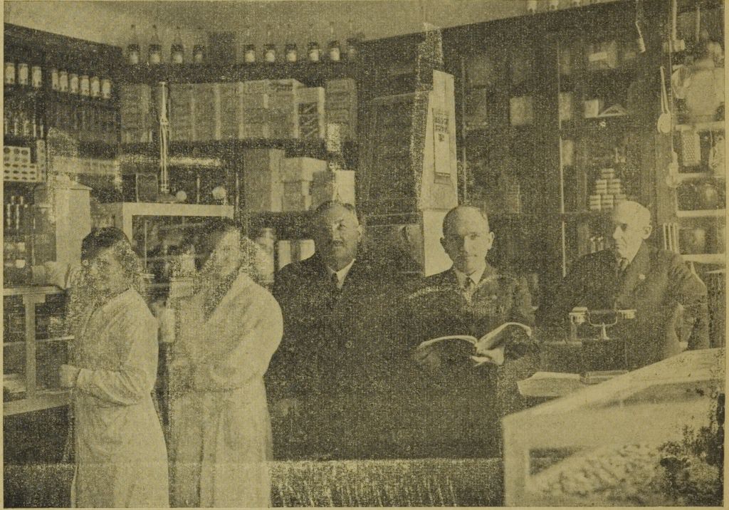 Wnętrze małego sklepu z półkami wypełnionymi towarami, na tle których pozują dwie kobiety w białych fartuchach i trzej mężczyźni w garniturach. Jeden z mężczyzn trzyma w rękach otwartą książkę. Na ladzie stoi telefon.