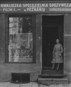 Kobieta stojąca na progu sklepu. Sklep jest na parterze kamienicy i prowadzą do niego 4 schodki. Widać okno z zaaranżowaną wystawą oferowanych towarów. Na górze szyld: Inwalidzka Spółdzielnia Spożywcza z ogr. odp. w Poznaniu, Filia nr 1, [ul.] Kraszewskiego 16