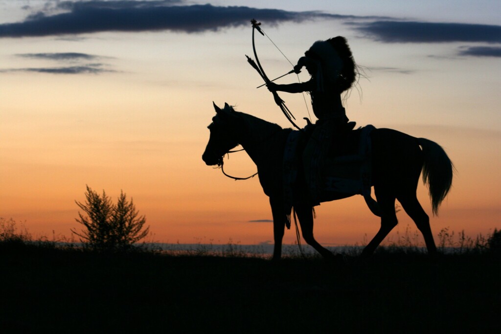 Zdjęcie Indianina z łukiem siedzącego na koniu, na tle zachodzącego słońca.