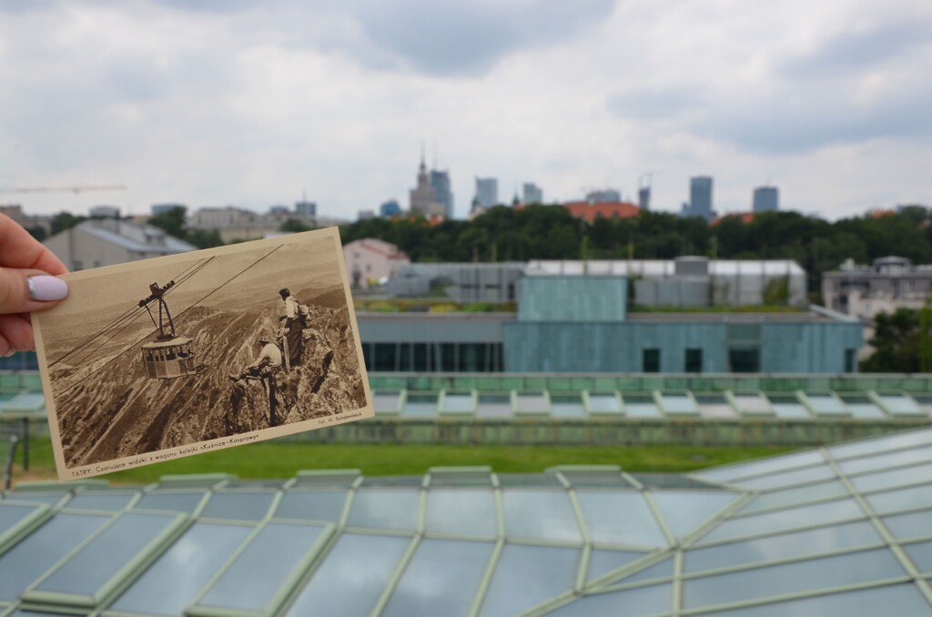 Zdjęcie, na pierwszym planie pocztówka z widokiem na wagonik kolejki linowej na tle gór, w tle dach BUW oraz budynki wydziału Filologii, a w oddali widoczne centrum Warszawy.