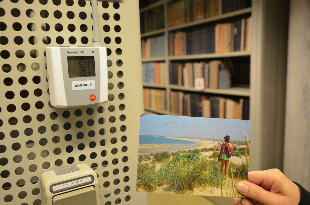 Na zdjęciu pocztówka z widokiem z wydm na plażę i morze, w tle magazyn biblioteczny i miernik temperatury wskazujący 20,3 st. Celsjusza
