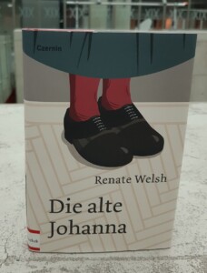 Zdjęcie okładki książki Die alte Johanna, na niej widoczny rysunek nóg w czerwonych skarpetach i czarnych butach na drewnianej podłodze.
