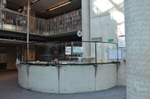 Zdjęcie betonowej, ustawionej w kształcie kręgu lady bibliotecznej z przeźroczystymi osłonami ustawionymi na niej. Za ladą dwie bibliotekarki.