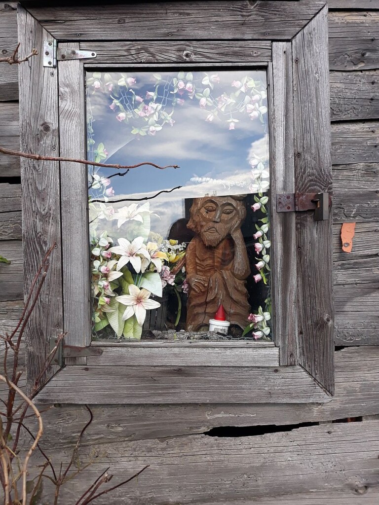 Zdjęcie przeszklonego okna starej chaty, widać ustawione w środku domu, na parapecie drewnianą figurkę oraz kwiaty.