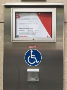 Zdjęcie informacji o godzinach otwarcia biblioteki, poniżej przycisk domofonu dla osób z niepełnosprawnościami.