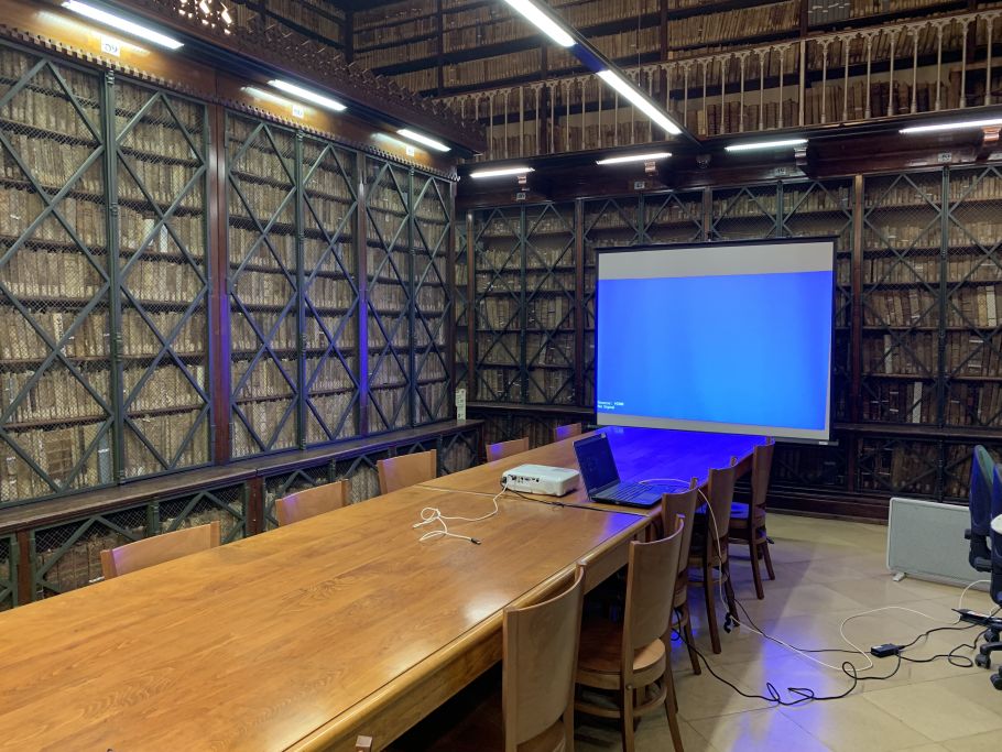 Zdjęcie czytelni bibliotecznej, z zakratowanymi regałami z książkami, stołem i ekranem do prezentacji.