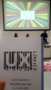 kartka z napisem UX Libsvi 06-08 06 2022 wpisanym w kod kreskowy. w tle ekran z tym samym logiem i ludzie siedzący przy stole konferencyjnym.