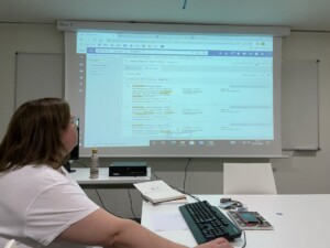 Zdjęcie pokazu, na pierwszym planie prelegentka, w tle ekran z otwartym oknem aplikacji bibliotecznej.