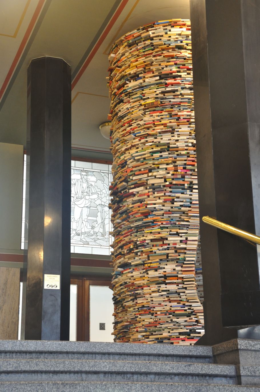 Zdjęcie wystającej zza filara wieży z książek, sięgającej od podłogi do sufitu.