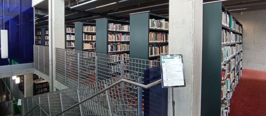 Tabliczka informacyjna ustawiona przy metalowych schodach, w tle regały z książkami i betonowe ściany.
