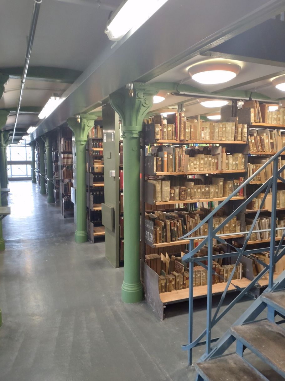 Zdjęcie magazynu bibliotecznego. Widać regały z książkami oraz zielone podpory stropu.